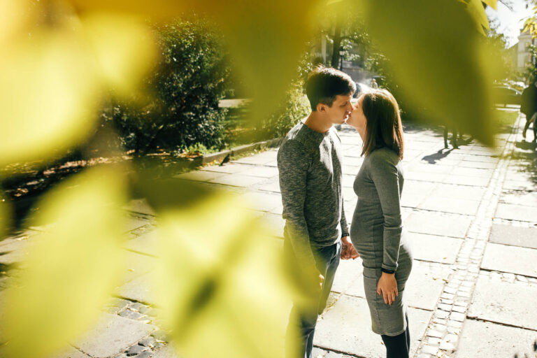 Couple standing in a tree-lined park pathway, leaning in for a kiss on a sunny day.