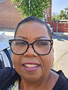 RealOptions Obria Medicical Clinic Staff. A close-up outdoor photo of a woman with dark hair pulled back, wearing black-rimmed glasses and smiling. A brick building and trees are visible in the background.