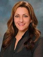 RealOptions Obria Medicical Clinic Staff. A headshot of a smiling woman with long, reddish-brown hair, wearing a dark, collared shirt against a blurred, gray background.