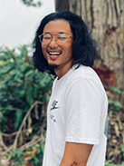 RealOptions Obria Medicical Clinic Staff. An outdoor headshot of a smiling man with long dark hair and glasses, wearing a white t-shirt.