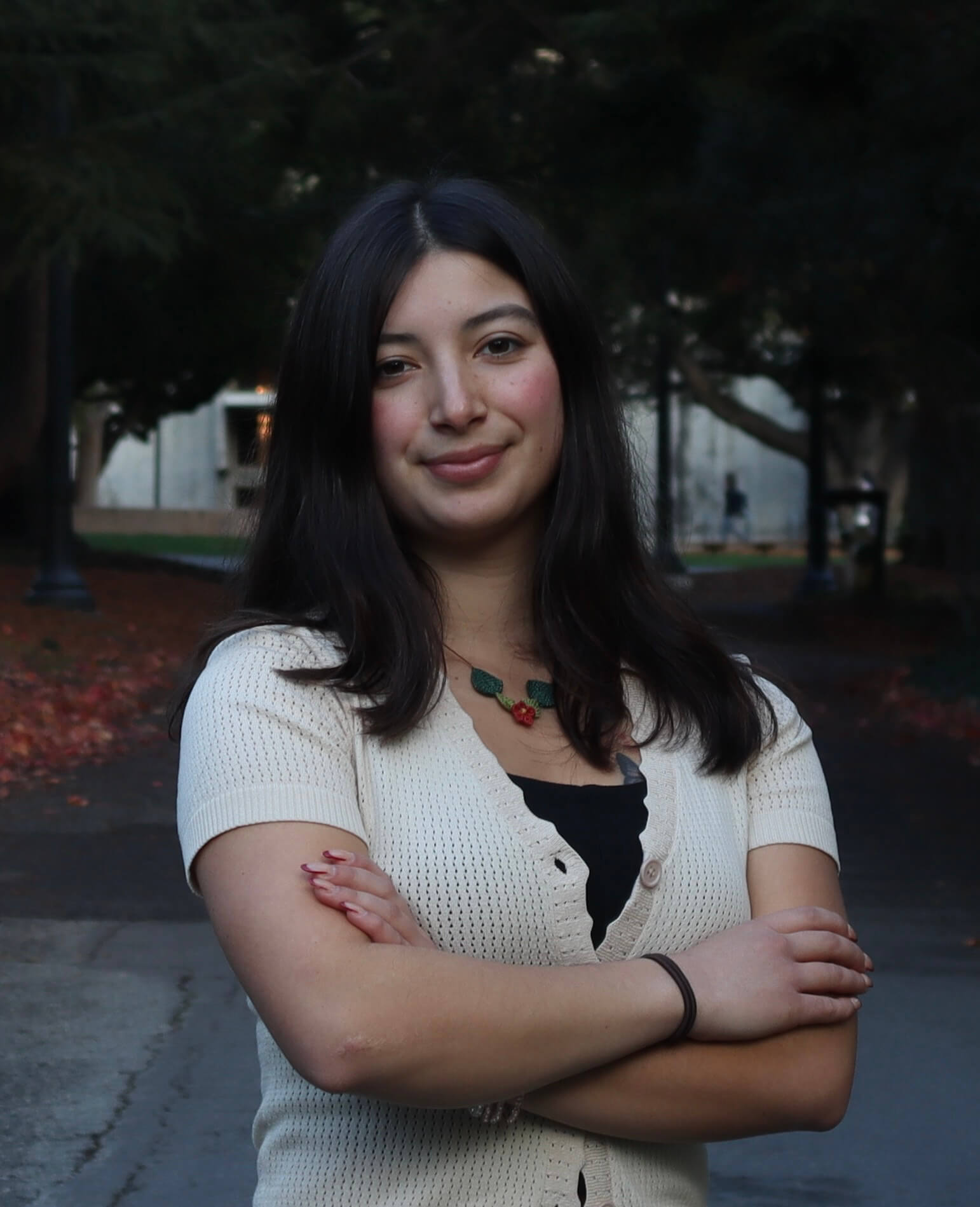 A young woman with long dark hair stands outdoors on a path, wearing a cream short-sleeved cardigan and a floral necklace, with her arms crossed and a slight smile. RealOptions Clinic - Education Staff.