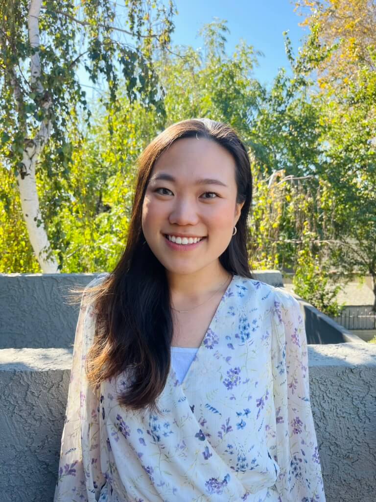 A smiling young woman with long dark hair, wearing a white floral-print wrap top, stands outdoors against a background of green trees and bright sunlight. RealOptions Clinic - Education Staff.